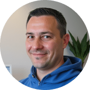 Person in blauem Pullover lächelnd vor grauer Innenwand mit Pflanze.